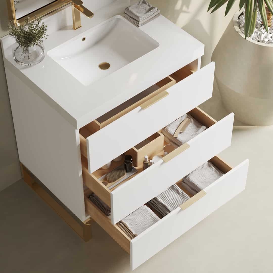 The Haldon Single Sink Bathroom Vanity in white boasts an integrated sink, gold fixtures, and three open drawers. The drawers are filled with neatly organized towels and toiletries. A plant and hand towel add a touch of nature on the side.