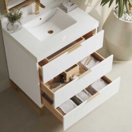 The Haldon Single Sink Bathroom Vanity in white boasts an integrated sink, gold fixtures, and three open drawers. The drawers are filled with neatly organized towels and toiletries. A plant and hand towel add a touch of nature on the side.