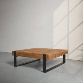 A wooden coffee table with a square top and black metal legs is positioned in a minimalist room with sunlight casting shadows on the wall.
