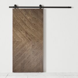 The Lucas 83" x 40" Reclaimed Wood Arrow Barn Door, featuring a herringbone pattern, is mounted on a black metal track against a plain white wall.
