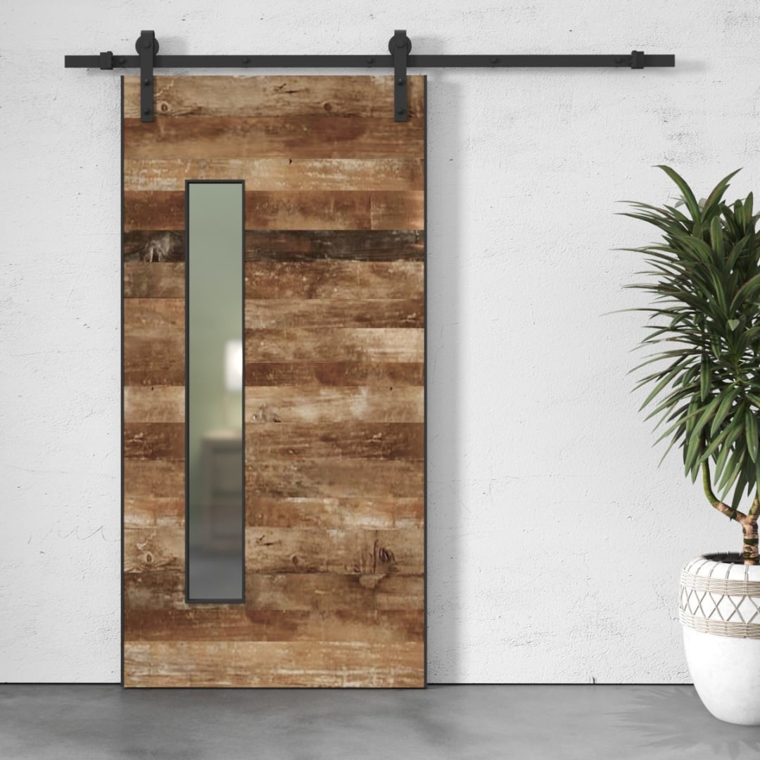 The Lucas 83" x 40" Reclaimed Wood Arrow Barn Door with a vertical glass panel is mounted on a black metal rail. A potted plant is placed to the right of the door against a white wall.