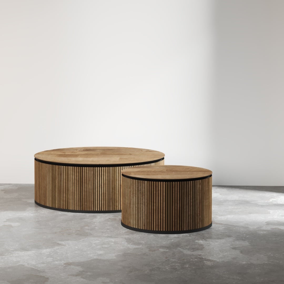 Two Bolivar Coffee Tables with slatted sides and round, wooden tops stand on a concrete floor against a plain, light wall. The tables are of different heights.