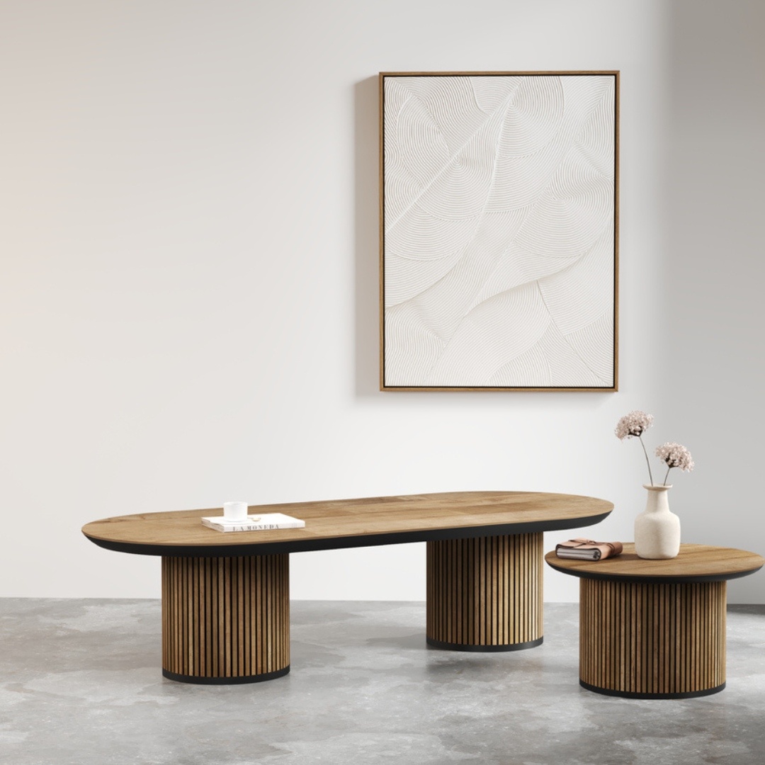 A minimalist room featuring the Oval Bolivar Coffee Table with a cylindrical base, accompanied by a matching side table. A white textured artwork adorns the wall behind them, while a vase filled with flowers and a cup elegantly rest on the tables.
