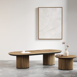 A minimalist room featuring the Oval Bolivar Coffee Table with a cylindrical base, accompanied by a matching side table. A white textured artwork adorns the wall behind them, while a vase filled with flowers and a cup elegantly rest on the tables.