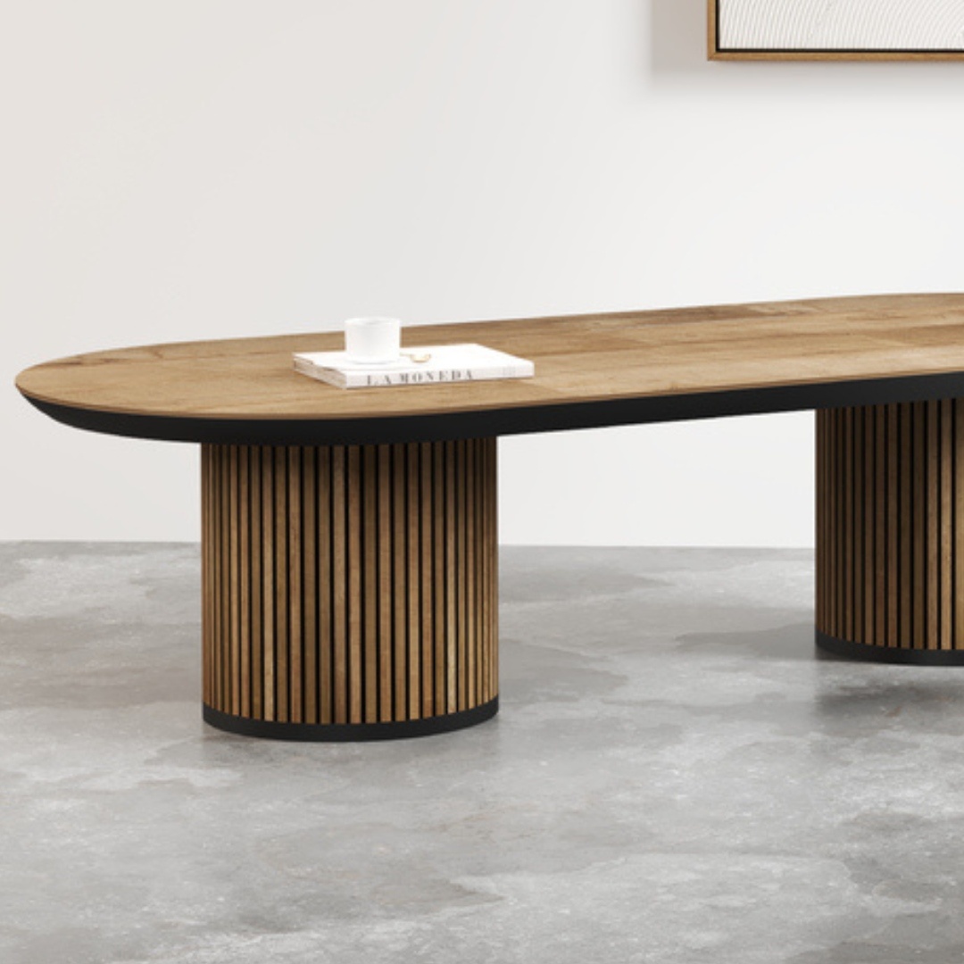 The Oval Bolivar Coffee Table, a modern wooden piece featuring rounded ends and ribbed cylindrical legs, is set against a minimalist background. A book and a small cup rest on the table.