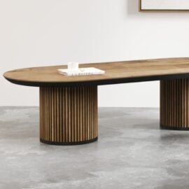The Oval Bolivar Coffee Table, a modern wooden piece featuring rounded ends and ribbed cylindrical legs, is set against a minimalist background. A book and a small cup rest on the table.