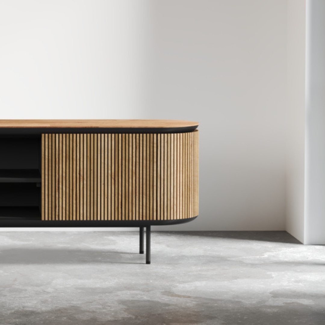 Bolivar 2 Shelf TV Stand featuring modern wooden design with vertical slats and black metal legs, placed in a minimalistic room with a concrete floor and white walls.