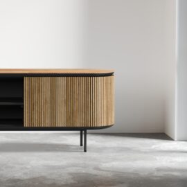 Bolivar 2 Shelf TV Stand featuring modern wooden design with vertical slats and black metal legs, placed in a minimalistic room with a concrete floor and white walls.
