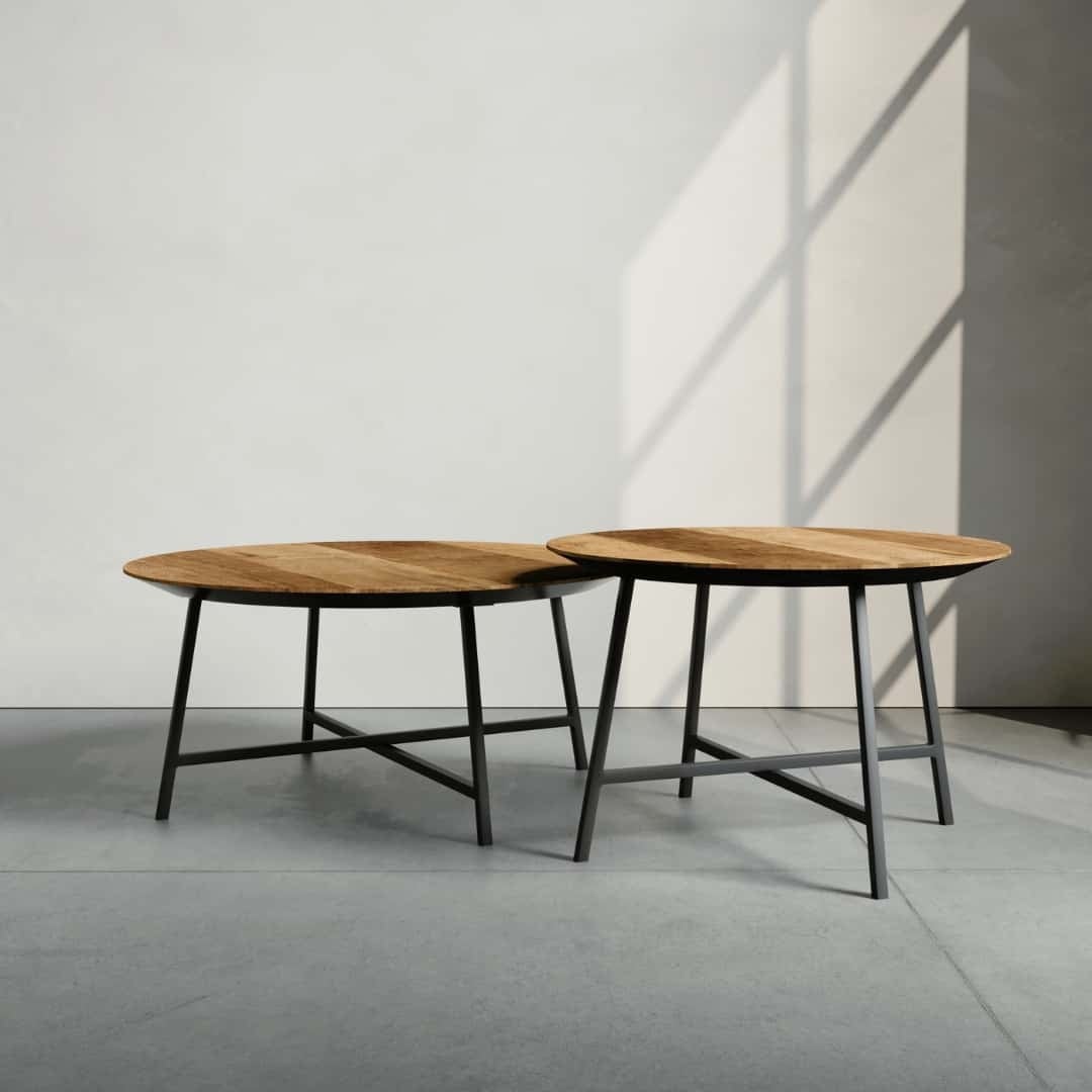 The 2 Piece Slim Round Coffee Table set, featuring wooden tops with black metal legs, is placed on a concrete floor in a well-lit room. Shadows cast by window panes are visible on the wall and floor.
