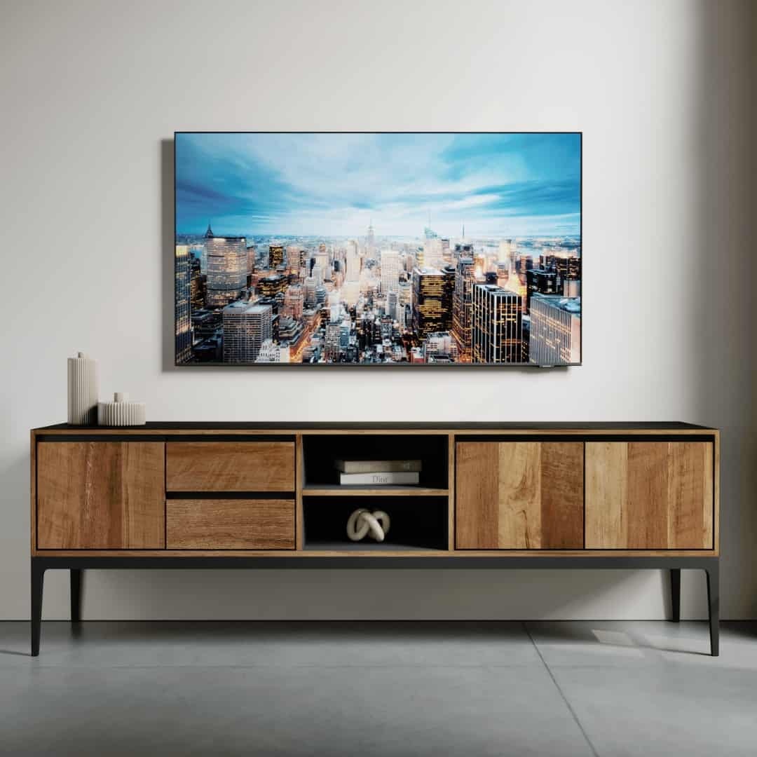 68 KOLT 2 SHELF TV STAND – 3 A Kolt 68" 2 Shelf TV Stand, holding several decorative items, sits below a flat-screen TV displaying a cityscape image. The TV is mounted on a white wall above the wooden console table, which features a black top and metal legs.