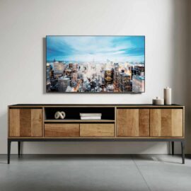 The Kolt 60" Wide TV Stand, featuring a modern wooden design with black legs, is placed against a white wall, adorned with decor items. Above it, a large flat-screen TV showcases a cityscape.