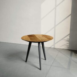 The Round Kolt Coffee Table, featuring a wooden surface and three black legs, stands on a gray floor near a wall with a window that casts shadows.