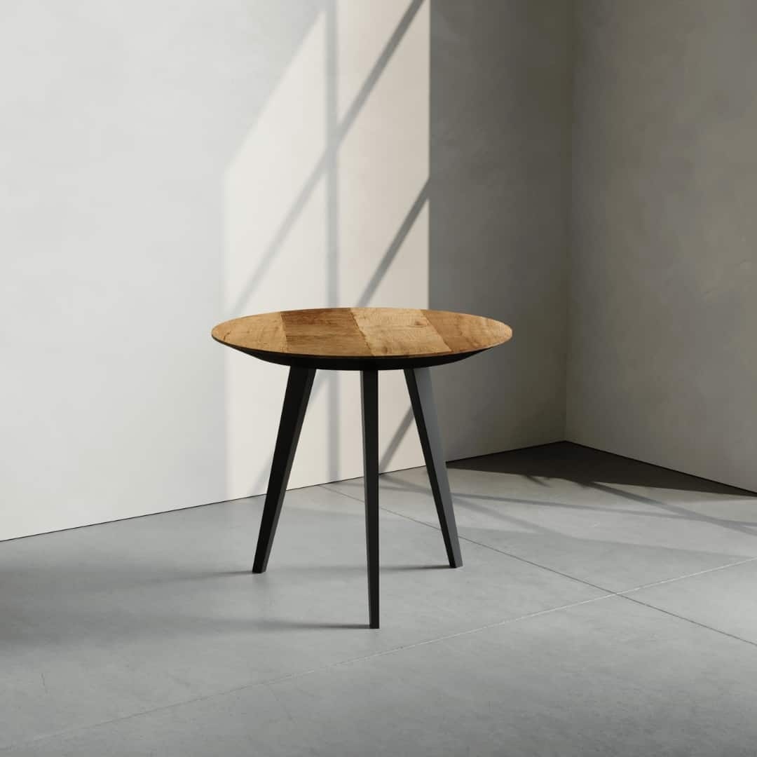 The Round Kolt Coffee Table, featuring a small round wooden top and sleek black legs, stands alone in a corner with natural light casting shadows on the walls and floor.