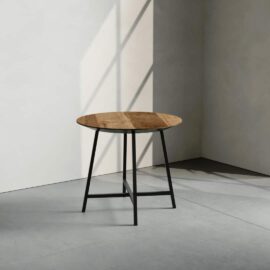 A Slim Round Coffee Table, featuring a wooden surface and black metal legs, is elegantly placed in a minimalist room adorned with concrete floors and walls. Sunlight streams in, casting intricate shadows on the floor.