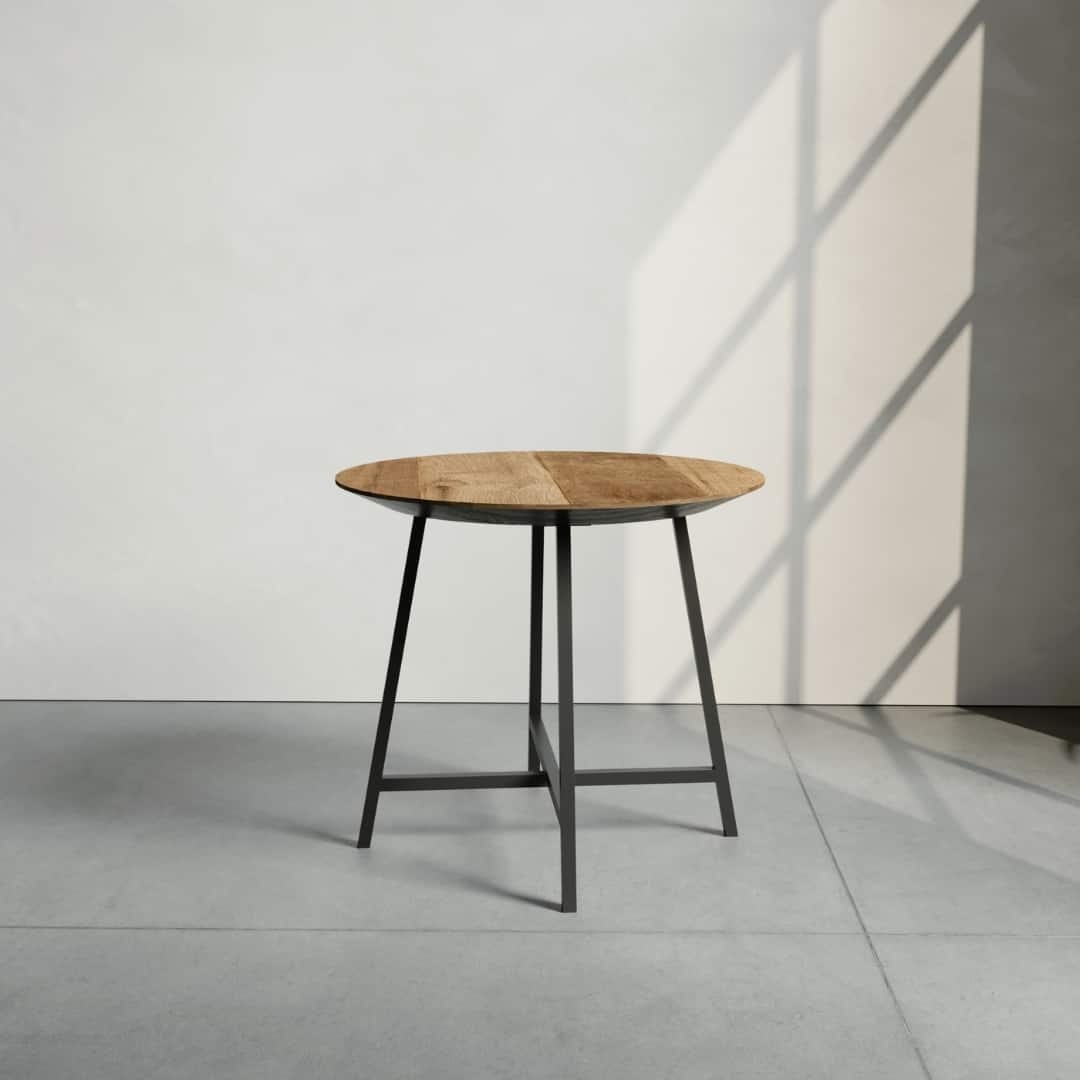 The Slim Round Coffee Table, featuring a circular wooden top and a black metal base, stands on a gray floor with sunlight casting a grid-like shadow on the wall behind it.