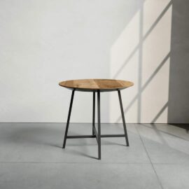 The Slim Round Coffee Table, featuring a circular wooden top and a black metal base, stands on a gray floor with sunlight casting a grid-like shadow on the wall behind it.