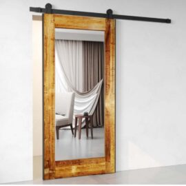 A Beveled Mirror Barn Door made of reclaimed wood is installed in an interior space, reflecting a room with a draped curtain and a small side table adorned with a decorative item.