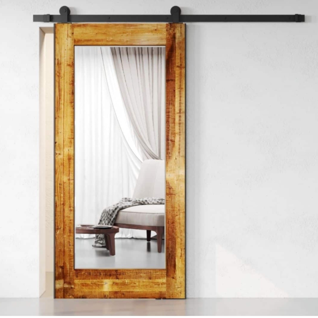 Beveled Mirror Barn Door (Reclaimed Wood) is partially open, showcasing its large mirror panel and revealing a minimalist room with white curtains and a beige chair.