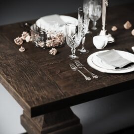 The Castello Dining Table - Café Noire is elegantly set with plates, cutlery, glasses, napkins, and various decorative items. The table setting features a white napkin, two forks, and an assortment of crystal glasses.