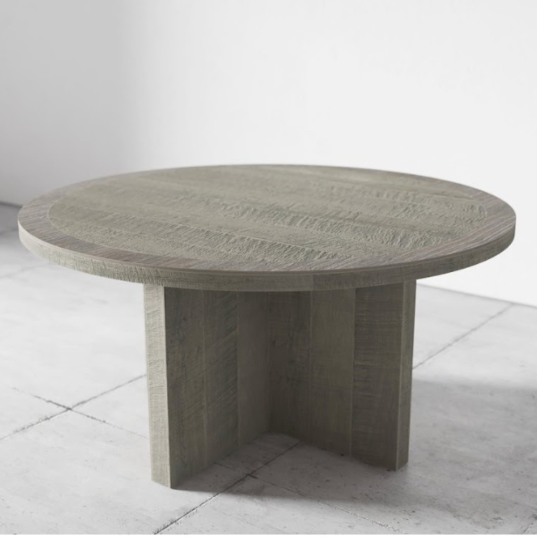 The Camden Round Dining Table - Rustic Grey, featuring a rustic finish and supported by a sturdy, cross-shaped pedestal base, stands on a concrete floor in a minimalist setting.