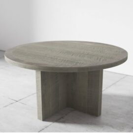 The Camden Round Dining Table - Rustic Grey, featuring a rustic finish and supported by a sturdy, cross-shaped pedestal base, stands on a concrete floor in a minimalist setting.