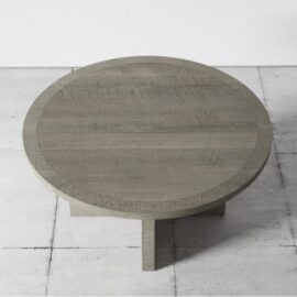 Placed on a tiled floor, the Camden Round Dining Table - Rustic Grey showcases a minimalist design with a round wooden surface in greyish-brown hues, supported by an elegant cross-shaped base.