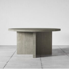 The Camden Round Dining Table in Rustic Grey, featuring a textured wooden surface and a geometric base, sits on a tiled floor against a plain white wall.