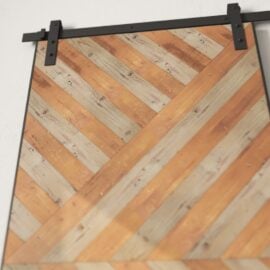 Enigma Reclaimed Wood Barn Door Grey with a chevron pattern made of alternating light and dark wooden strips, mounted on black sliding hardware against a white wall.