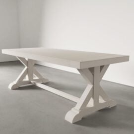The Madera Dining Table - Pure White, a large rectangular wooden piece with X-shaped legs and a crossbar, stands on a gray concrete floor in a minimalist room.