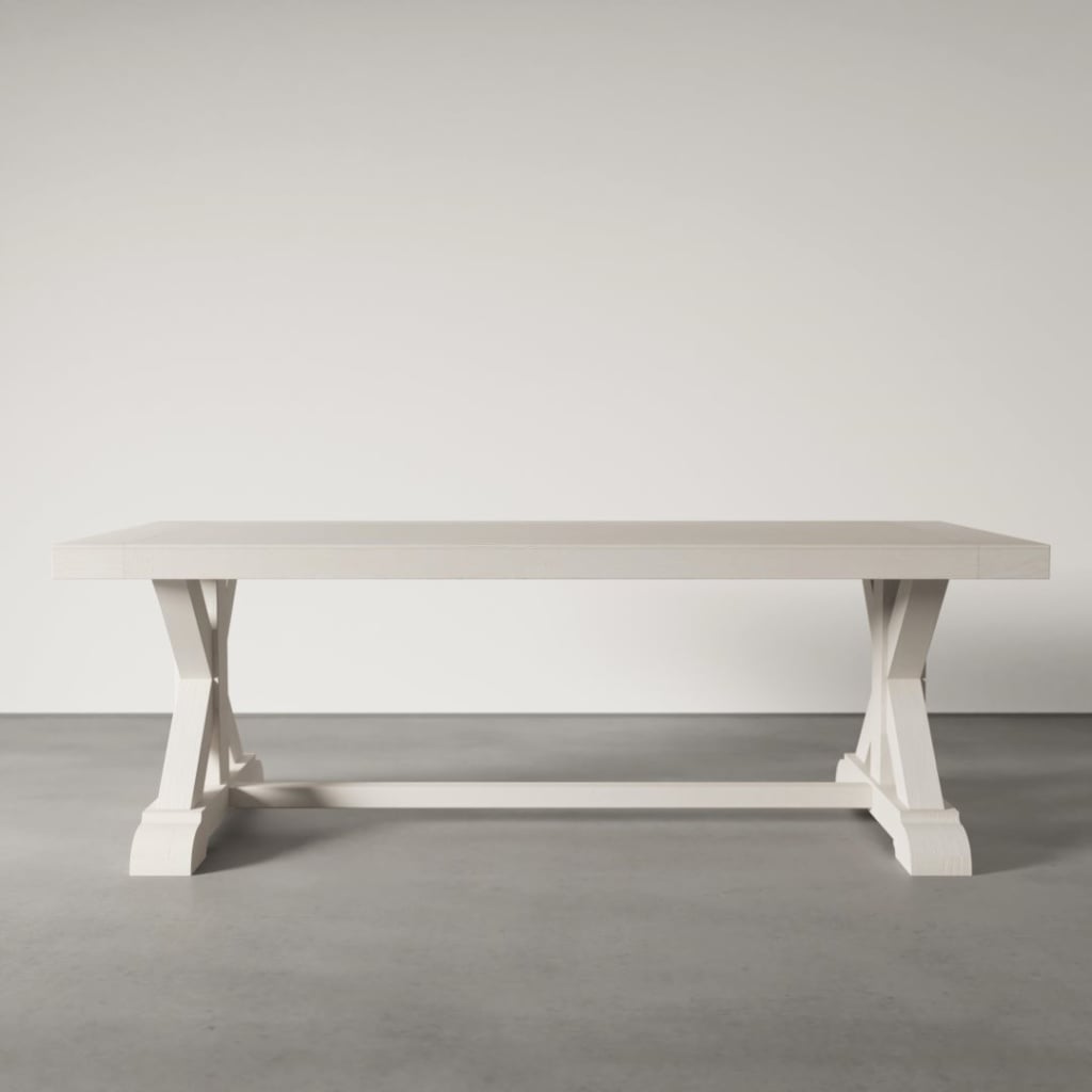 The Madera Dining Table - Pure White, featuring a rectangular design and an X-shaped base, stands on a gray concrete floor against a plain light-colored wall.