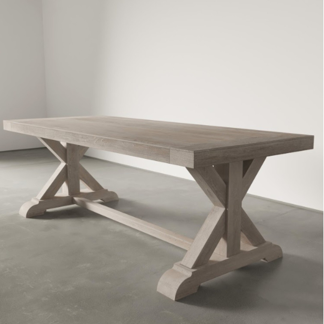 A spacious, rectangular Madera Dining Table - White Wash with an X-shaped trestle base is set on a concrete floor, framed by a plain white backdrop.