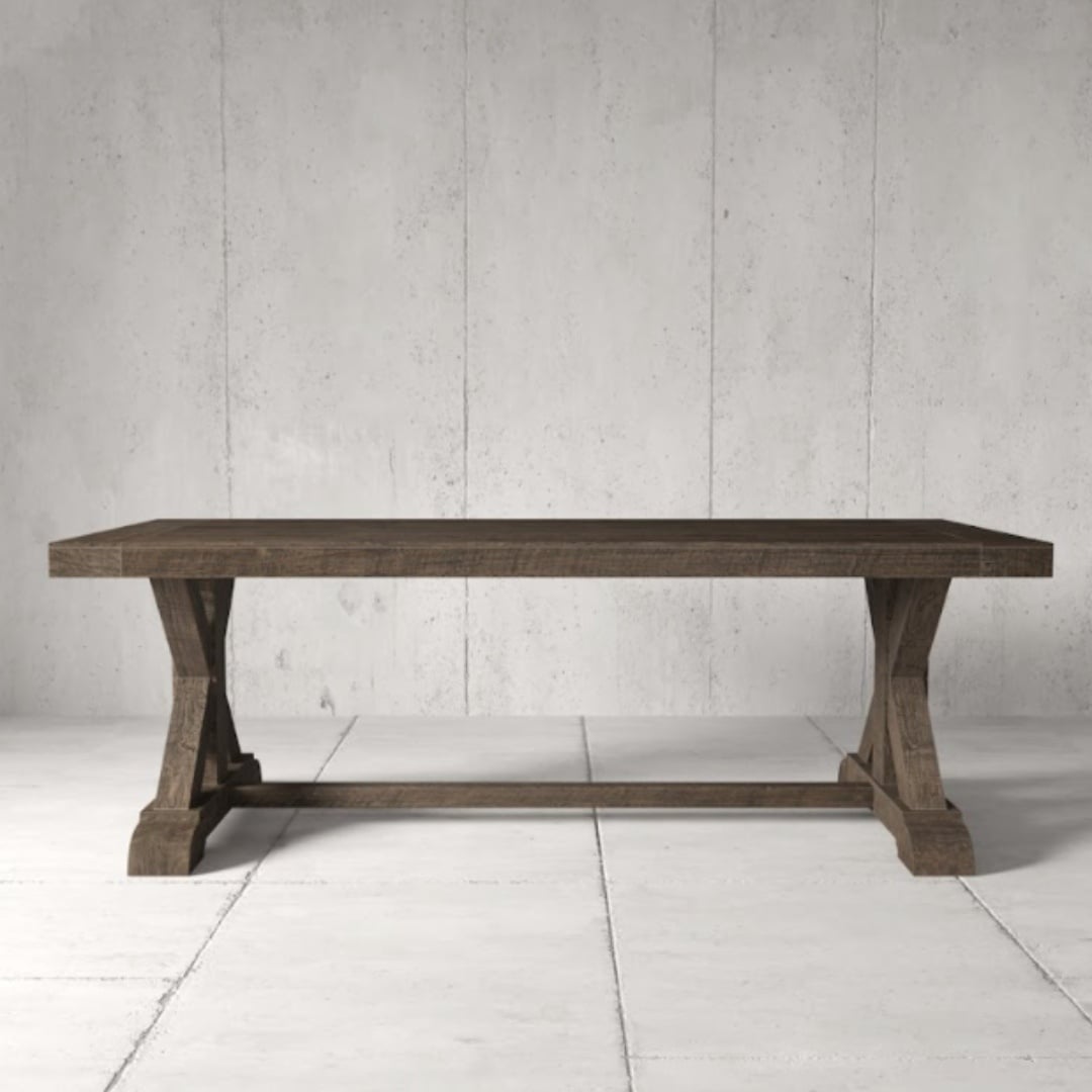 The Madera Dining Table - Café Noire, featuring sturdy, carved wooden legs, stands on a light, tiled floor against a concrete wall.