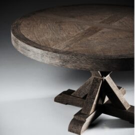The Madera Round Table, reminiscent of a Café Noire setting, features a detailed carved surface and a sturdy, ornate pedestal base on a light grey background.