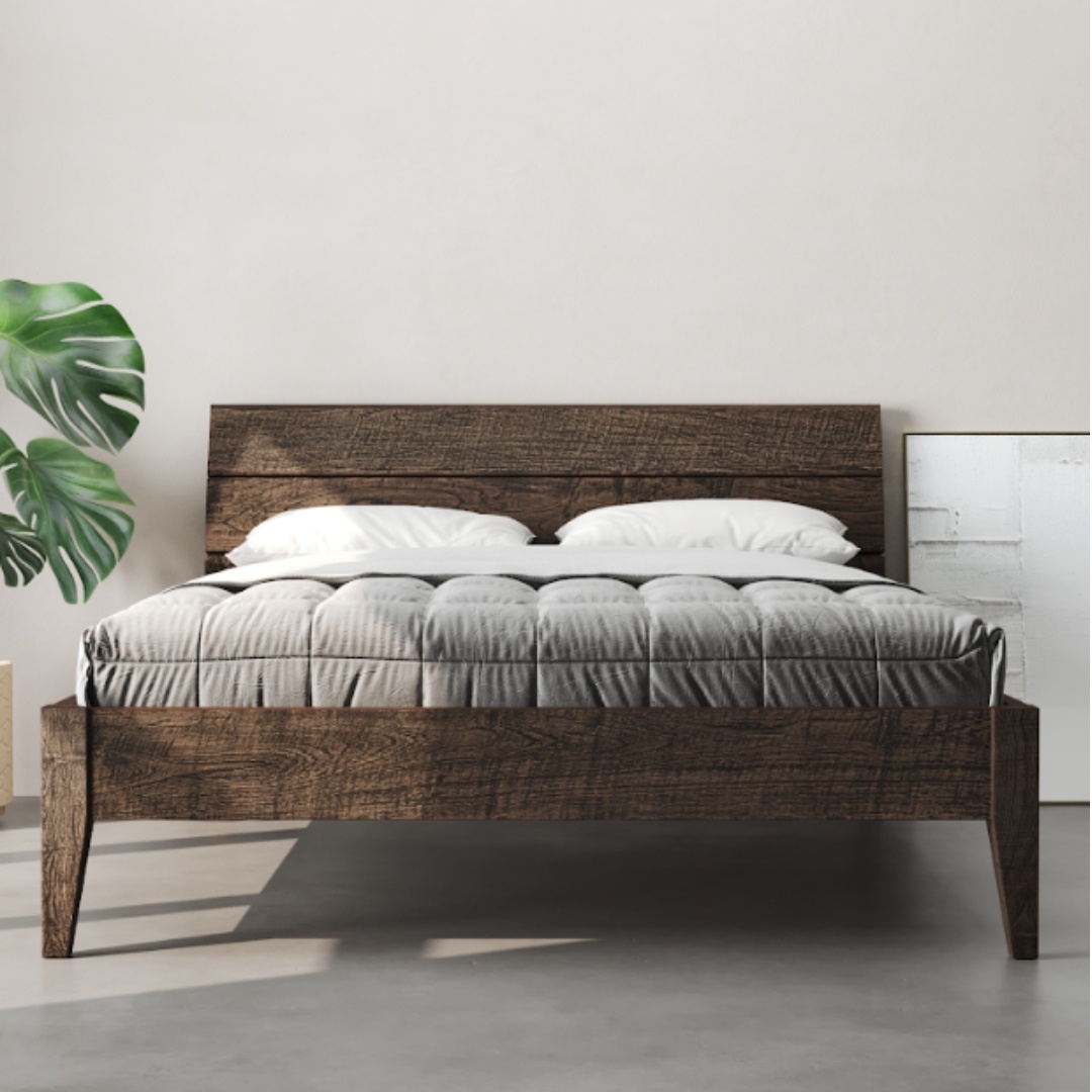 A Tuscan Bed (Café Noire) frame with a grey quilted mattress and two white pillows, placed against a white wall, with a green leafy plant on the left.