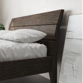 The Tuscan Bed (Café Noire) features a wooden frame and is adorned with white pillows and bedding. Beside it, a framed artwork with white textured paint leans against the wall.