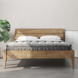 The Tuscan Bed (Natural) features a grey quilted mattress and white pillows, with a plant positioned to the left and a white painting leaning against the wall on the right.