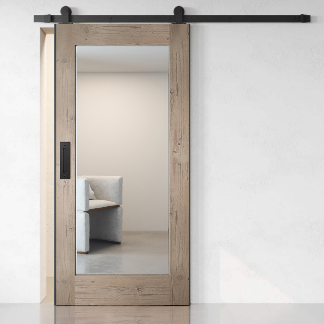A wooden sliding barn door with a large glass pane, partially open, reveals a minimalistic room with a light-colored armchair inside. The rustic grey tone of the door adds an element of charm to the setting.