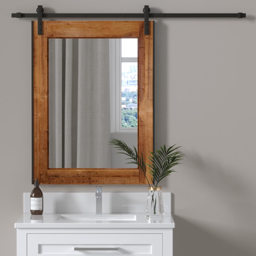 A bathroom vanity features a Reclaimed Wood Rectangular Sliding Vanity Mirror, complemented by a white sink with a silver faucet. The setup includes a brown soap dispenser and a glass vase filled with green plant leaves, with a window in the background.