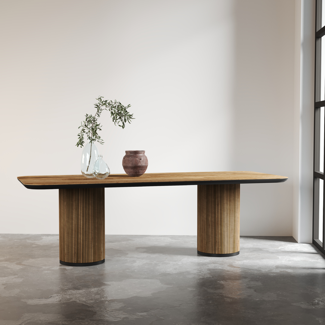 The Bolivar Dining Table, featuring a modern wooden design with two cylindrical legs, is adorned with two clear vases holding green branches and a small brown ceramic pot, all set in a bright room with a concrete floor and large window.