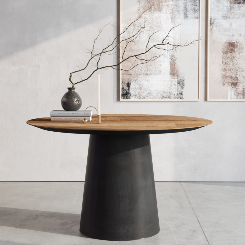 The Atelier Round Dining Table featuring a wooden top and black base showcases a vase with branches, neatly stacked books, and a candle, set against a minimalist wall adorned with abstract artwork.
