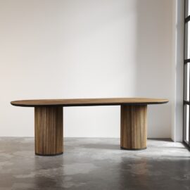 The Bolivar Oval Dining Table, featuring a vertical slat design on cylindrical legs, stands on a concrete floor in a minimalist room bathed in natural light.