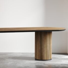 The Bolivar Oval Dining Table boasts a contemporary wooden design with an oval-shaped top and a cylindrical base accented by vertical grooves. It is placed against a minimalist white wall on a concrete floor.