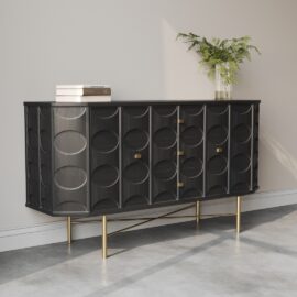 The Alba 60" Side Cabinet showcases black circular patterns, accented by gold handles and legs, standing gracefully against a light wall. A potted fern and a stack of books embellish the top, while the room's concrete floor enhances its modern appeal.