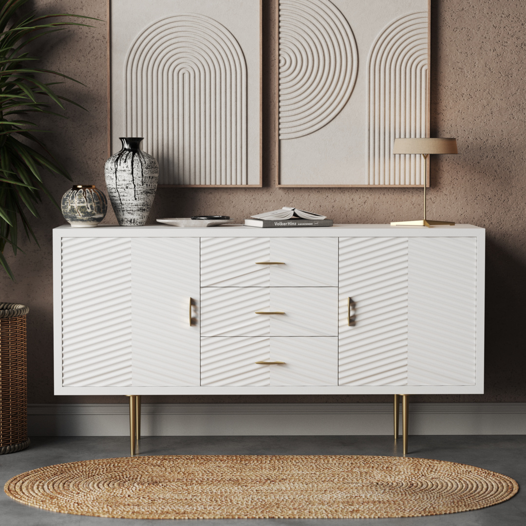The Arista 60" Side Cabinet (White Wash) showcases a white wash finish with a textured pattern, complemented by gold handles and legs. Its spacious 60-inch surface is adorned with decorative vases, books, and a lamp. Two abstract artwork pieces are displayed above it, while a round woven rug adds warmth to the floor below.