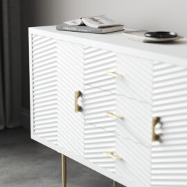 The Arista 60" Side Cabinet in White Wash boasts textured doors and sophisticated gold handles, elegantly perched on slender legs. Offering ample space for decor such as books and dishes on its smooth top, it creates a stunning contrast against the gray concrete floor.