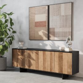 The Atelier Dining Buffet with wooden doors stands against a white wall, topped with decorative items, next to a green potted plant. Two framed abstract artworks are mounted above it.