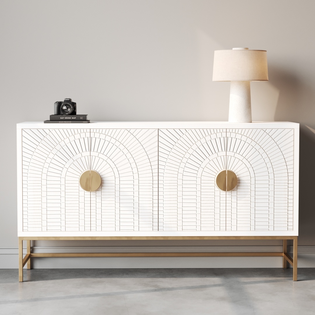 The Coral 60" Side Cabinet features a white finish with decorative geometric patterns, round gold handles, and a gold base. It is styled with a lamp and a camera on top, set against a plain light gray wall, adding a subtle coral accent to the space.