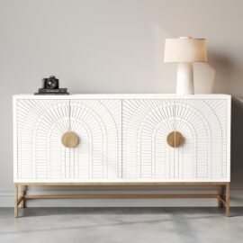 The Coral 60" Side Cabinet features a white finish with decorative geometric patterns, round gold handles, and a gold base. It is styled with a lamp and a camera on top, set against a plain light gray wall, adding a subtle coral accent to the space.