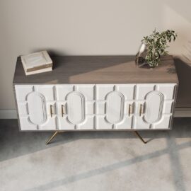 The Lugo 60" Side Cabinet features a contemporary design with a wooden top, white geometric-patterned doors, elegant gold handles, and decorative accents—a plant in a glass vase on the right side and a book stacked on the left side.