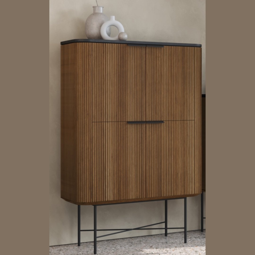 The Osaka Tall Display Cabinet, featuring vertical grooves and supported by black metal legs, stands against a beige wall. Decorative white vases adorn its top.